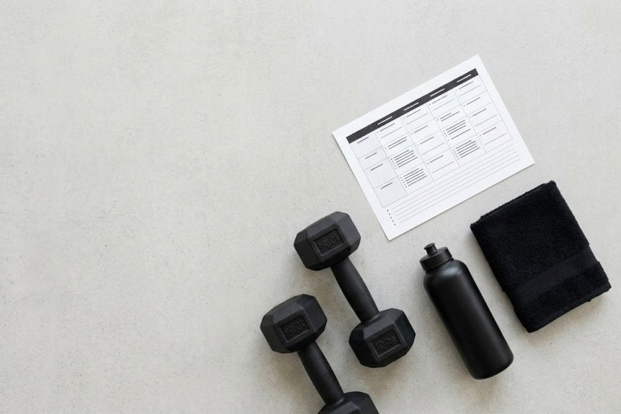 Trainingsplan auf Papier, daneben Hanteln, Trinkflasche und Handtuch auf hellem Beton